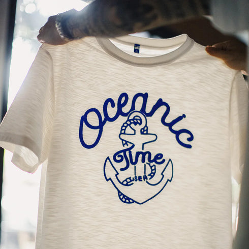 White t-shirt with blue 'Oceanic Time' anchor design held by a person.